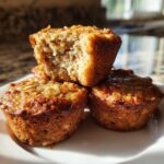 Close-up of three golden brown oatmeal muffins stacked on a white plate, one broken open showing the moist interior.