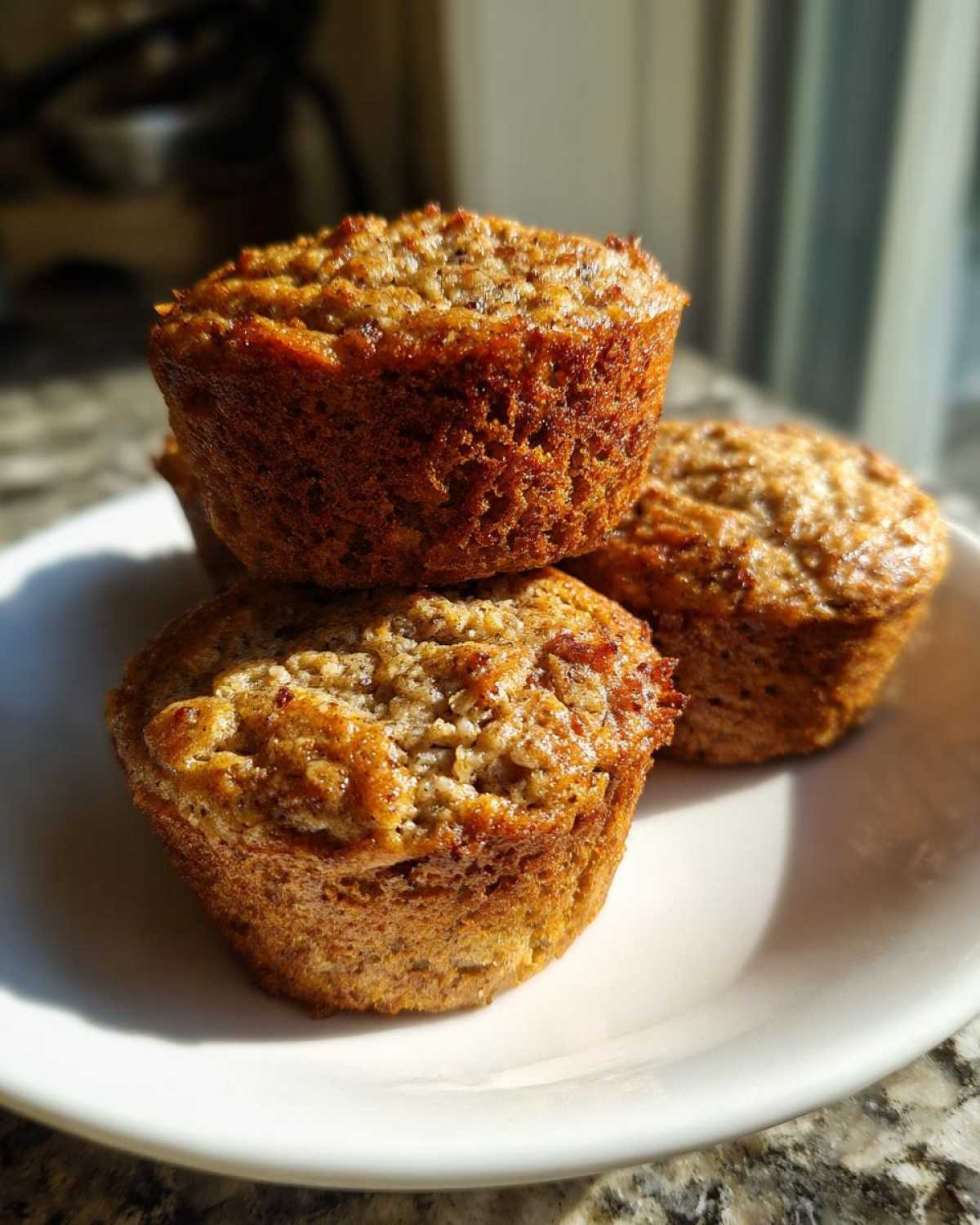 A stack of three golden brown, moist oatmeal muffins sitting on a white plate, highlighted by sunlight.