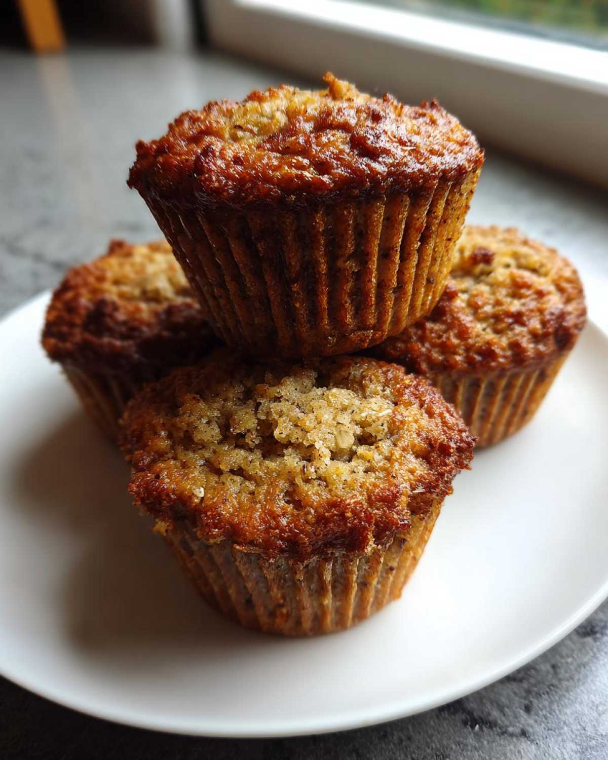 A stack of four freshly baked, moist oatmeal muffins with deeply golden brown, slightly crunchy tops, resting on a white plate.