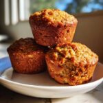Three golden-brown, moist oatmeal muffins stacked on a white plate, one with a bite taken out.