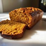 A close-up of a moist pumpkin bread loaf, with one thick slice cut and resting beside it on a white marble surface.