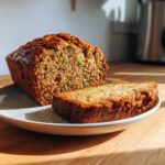 A golden-brown loaf of zucchini banana bread, partially sliced, resting on a white plate.