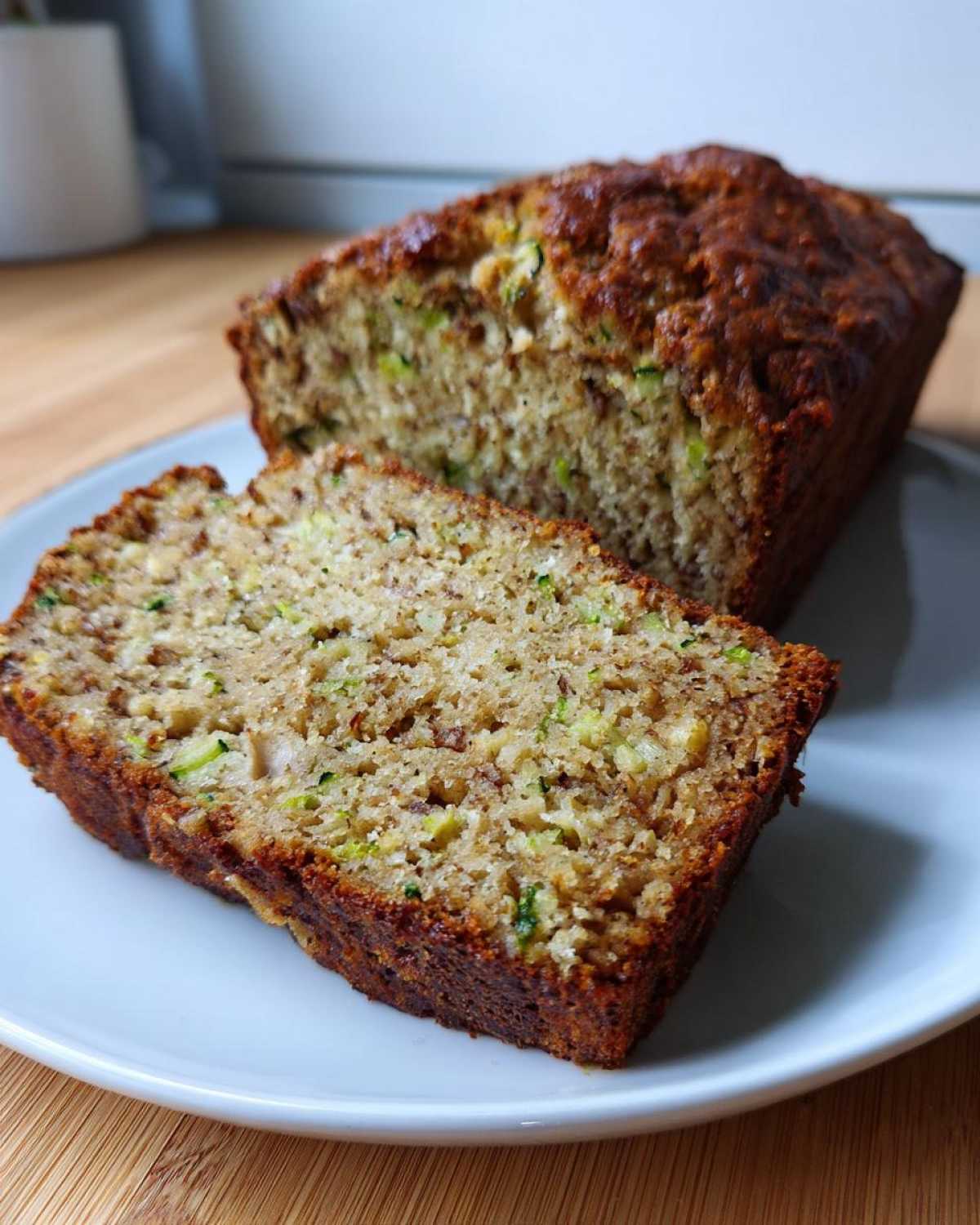 A thick slice of moist zucchini banana bread resting on a white plate next to the rest of the loaf.
