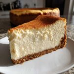 A thick, creamy slice of new york cheesecake with a golden-brown top and graham cracker crust on a white plate.