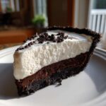 A close-up slice of no bake Oreo pie showing a dark crust, chocolate filling, and thick white cream topping sprinkled with crumbs.