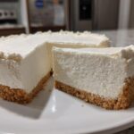 A slice cut from a whole, creamy white Nobake Cheesecake Silky Sets Perfectly, showing a thick graham cracker crust.