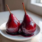 Two vibrant, deep red poached pears glistening with syrup, served on a white plate.
