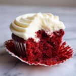 Close-up of a red velvet cupcakes with a bite taken out, revealing the moist red cake and thick cream cheese frosting.