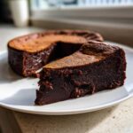 A dense, fudgy slice of flourless chocolate cake dusted with cocoa powder, with the rest of the cake in the background.