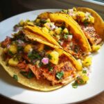 Three delicious Salmon Tacos with Pineapple Salsa piled high on a white plate, featuring flaky salmon and bright yellow pineapple chunks.