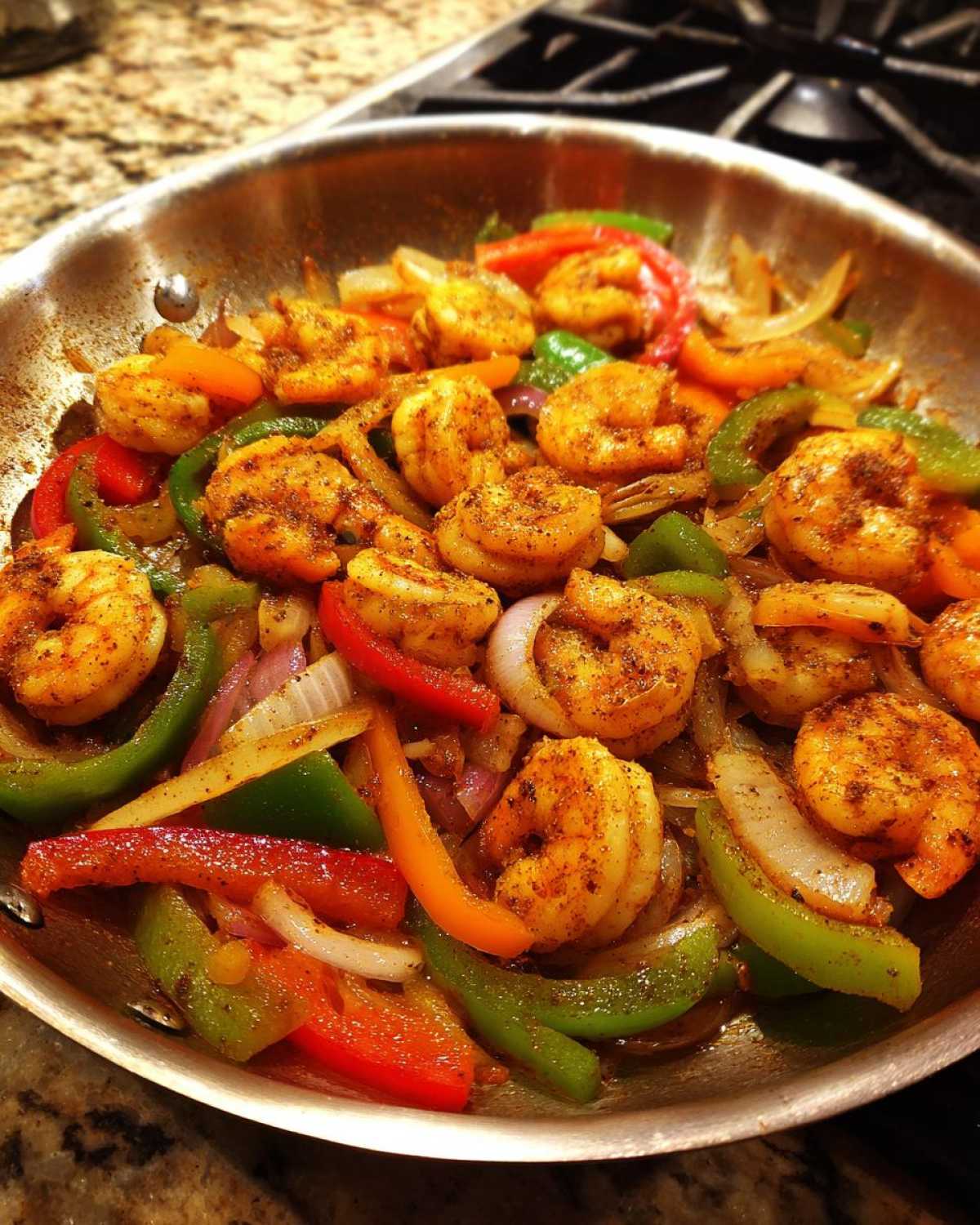 Close-up of seasoned shrimp cooking with sliced bell peppers and onions for shrimp fajitas in a stainless steel skillet.