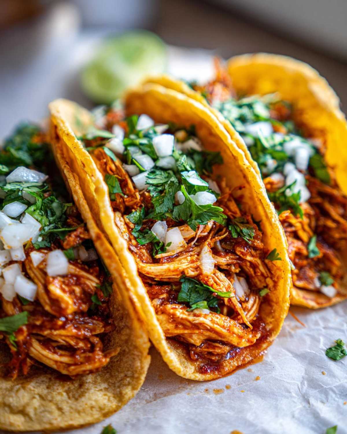 Close-up of three corn tortilla chicken tacos filled with shredded chicken in sauce, topped with diced white onion and fresh cilantro.