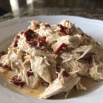 A close-up of shredded crockpot marry me chicken coated in a creamy sauce with pieces of sun-dried tomatoes.