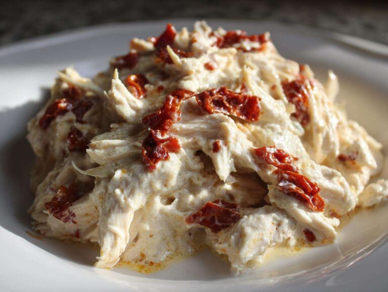 A close-up of shredded crockpot marry me chicken coated in a creamy sauce and topped with sun-dried tomatoes.