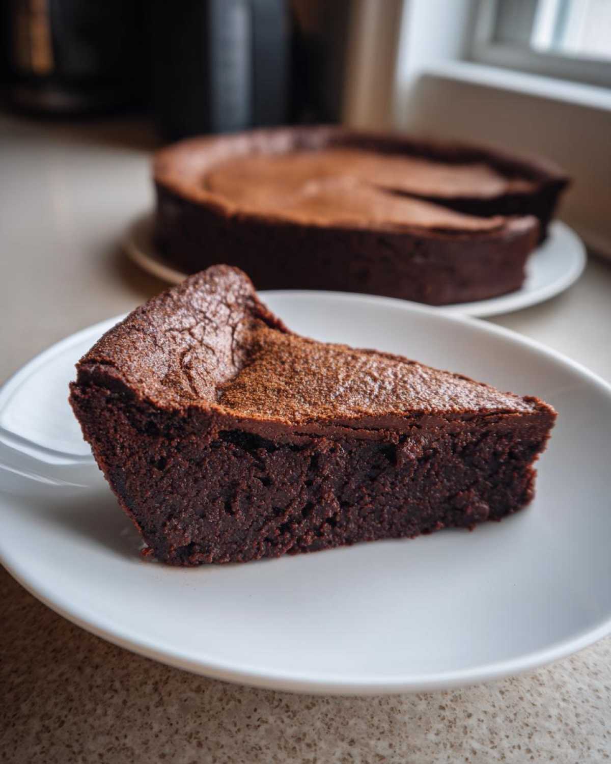 A thick slice of rich, dark flourless chocolate cake dusted with cocoa powder, served on a white plate.