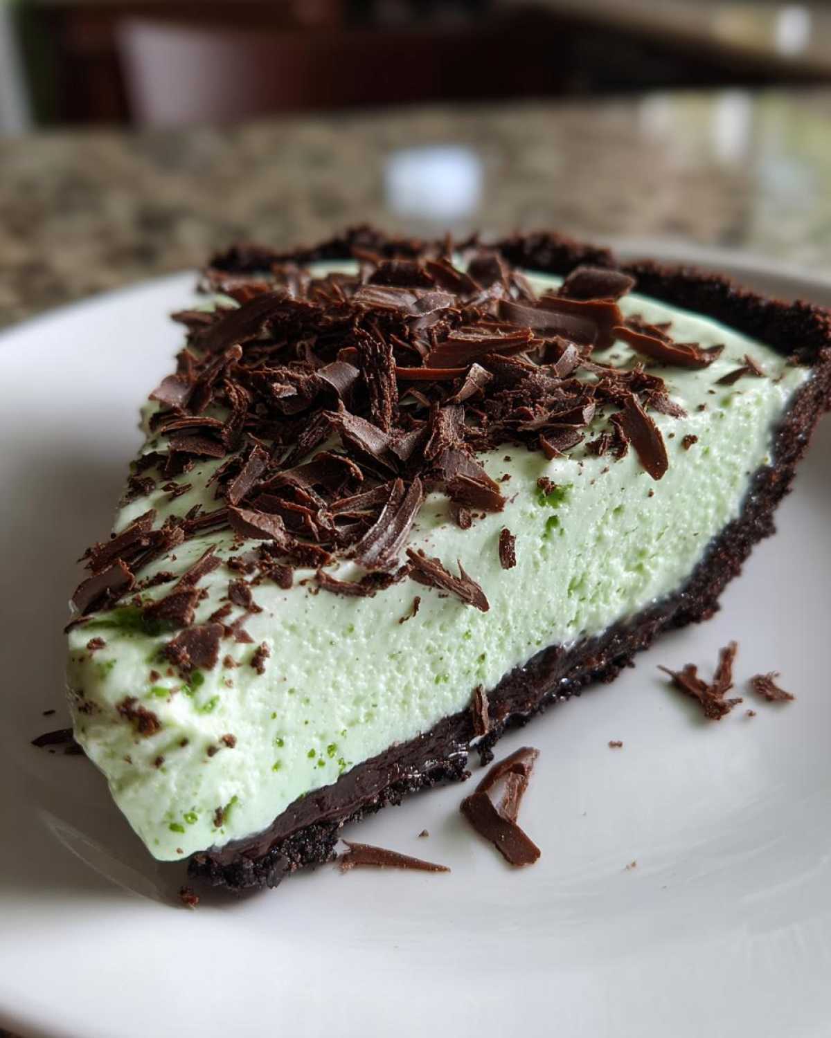 A close-up of a creamy, light green slice of grasshopper pie with a dark chocolate crust, topped with chocolate shavings.