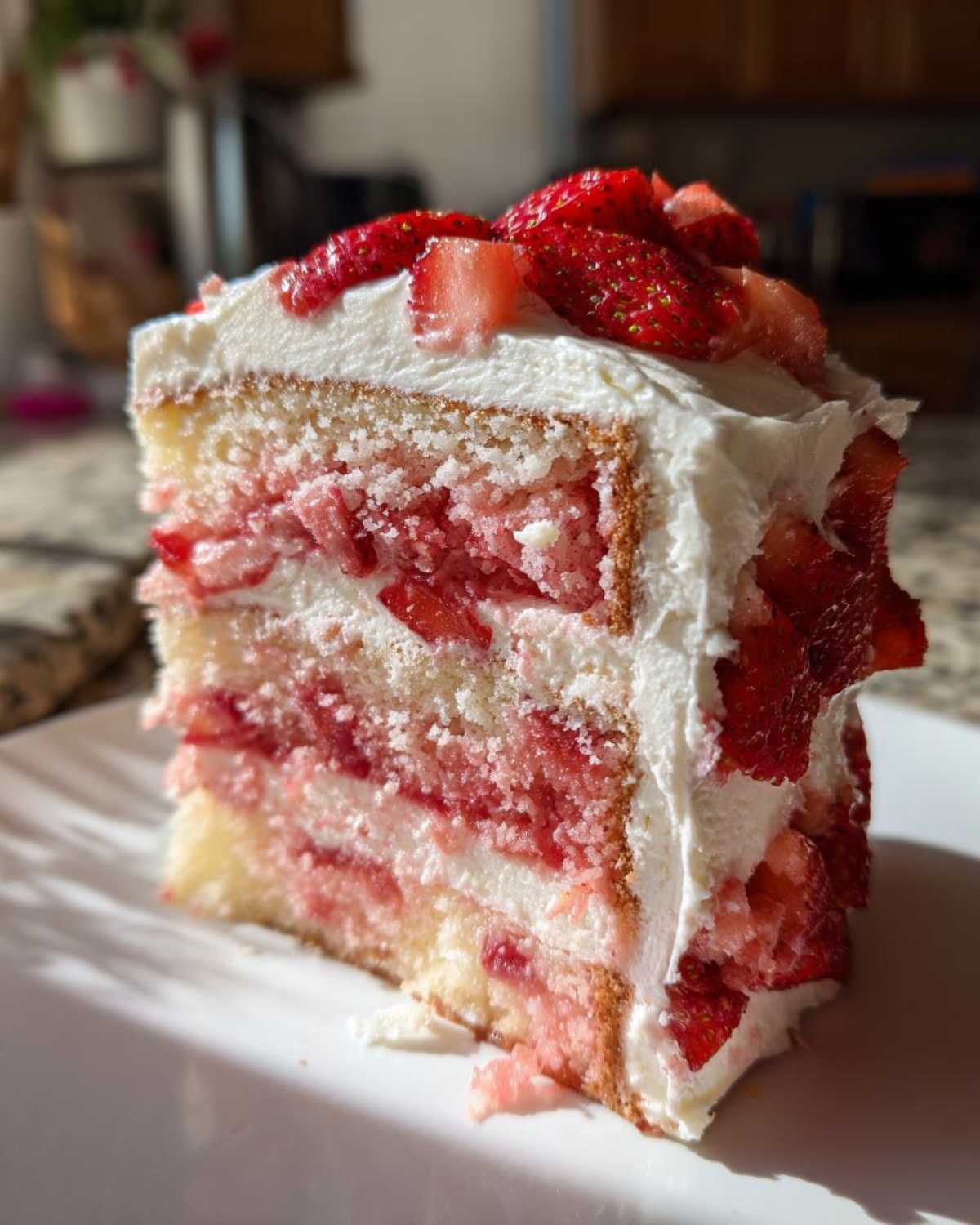 A tall slice of layered strawberry cake with white frosting and fresh strawberries on top.