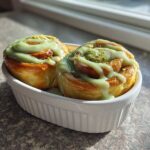Two Small Batch Pistachio Cream Cinnamon Rolls drizzled with green icing and topped with crushed nuts in a white ramekin.