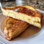 Two stacked slices of golden brown sourdough french toast drizzled with syrup on a white plate.