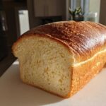 A freshly baked loaf of soft sourdough sandwich bread showing a golden, shiny crust and a tender crumb structure.