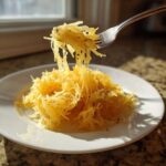 A fork lifts strands of cooked spaghetti squash recipe from a white plate, showing its noodle-like texture.