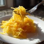 A fork lifting bright yellow, cooked spaghetti squash recipe strands from a white plate.