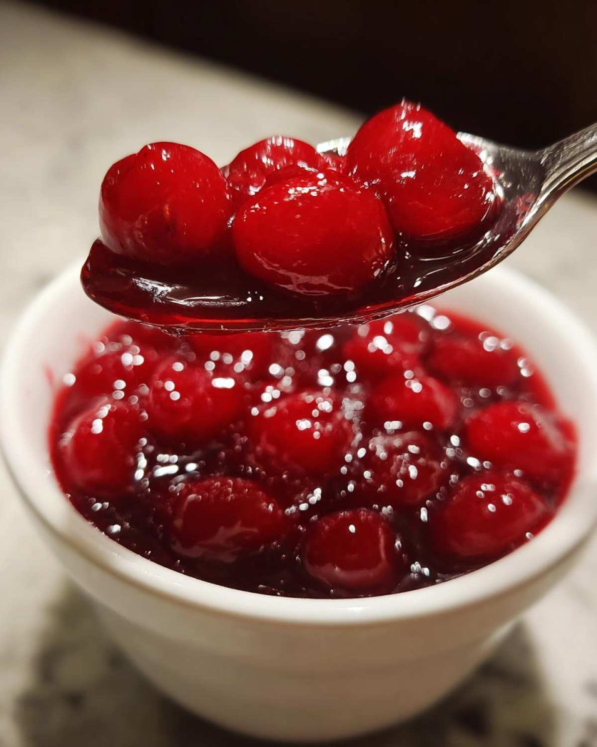 A spoonful of glossy, bright red cherry pie filling being lifted above a white bowl full of the same cherry pie filling.