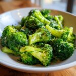 Close-up of bright green steamed broccoli florets tossed with herbs and seasoning in a white bowl.