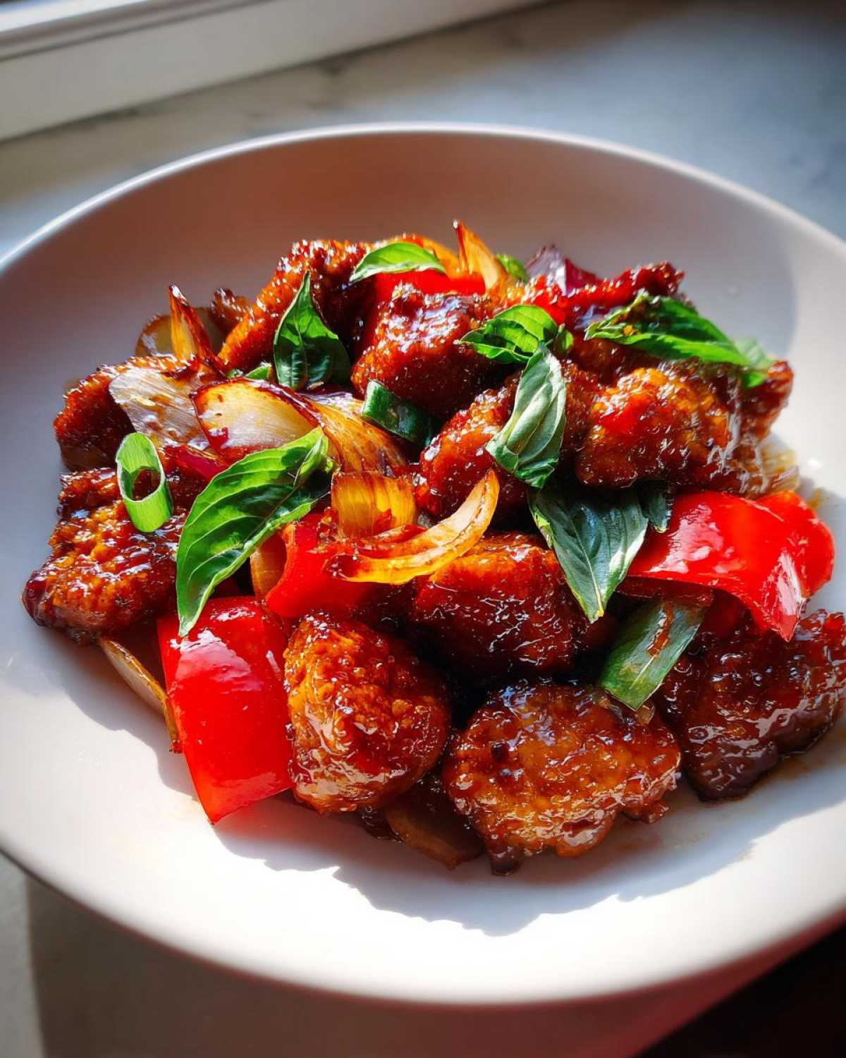 Close-up of glistening Sweet Spicy Thai Chicken pieces mixed with red bell peppers, onions, and fresh basil leaves.