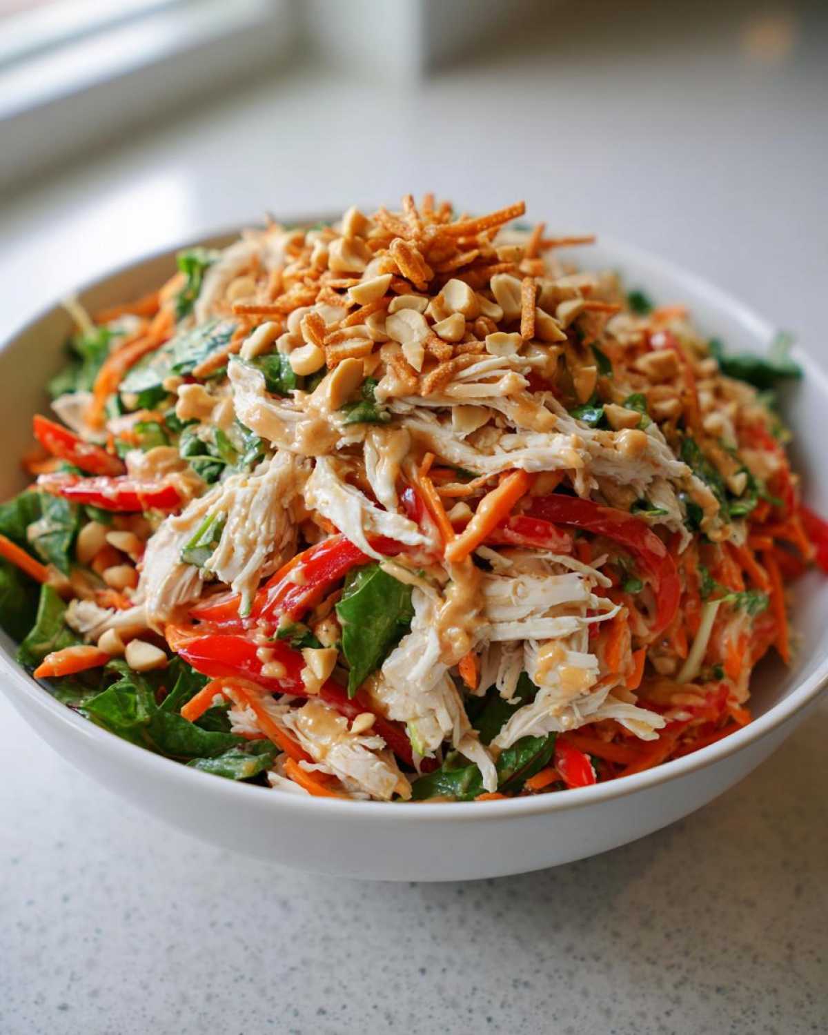 A vibrant bowl of thai chicken salad featuring shredded chicken, carrots, red peppers, greens, and topped with peanut dressing and crunchy peanuts.
