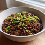 A white bowl filled with rich, dark Vegan Korean Bulgogi Lentils, garnished with sliced green onions and sesame seeds.