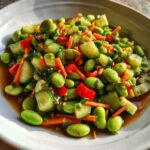 Close-up of a bright, colorful edamame salad featuring shelled edamame, cucumber chunks, shredded carrots, and red peppers in a savory dressing.