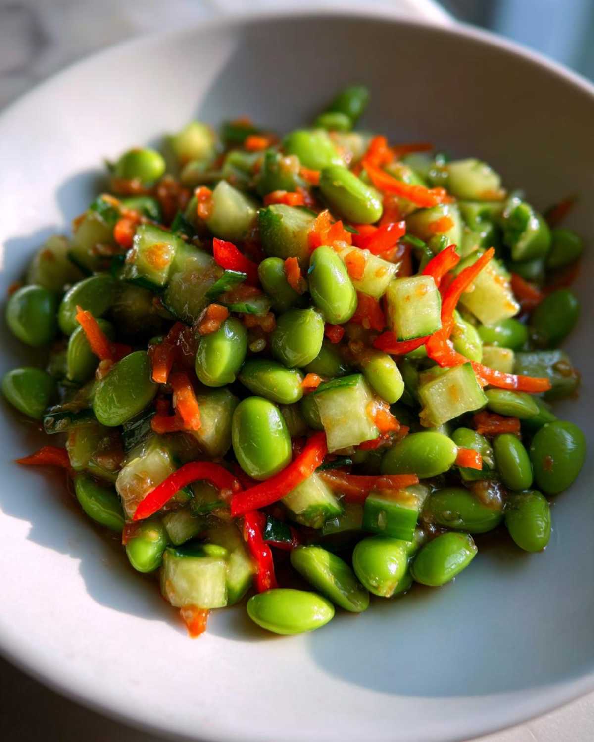 Close-up of a bright green edamame salad mixed with diced cucumber, shredded carrots, and strips of red pepper.