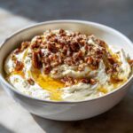 A close-up of creamy whipped goat cheese topped with glistening honey and toasted pecans in a white bowl.
