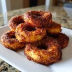 A stack of golden Air Fryer Cinnamon Apple Rings heavily coated in cinnamon sugar, served on a white rectangular plate.