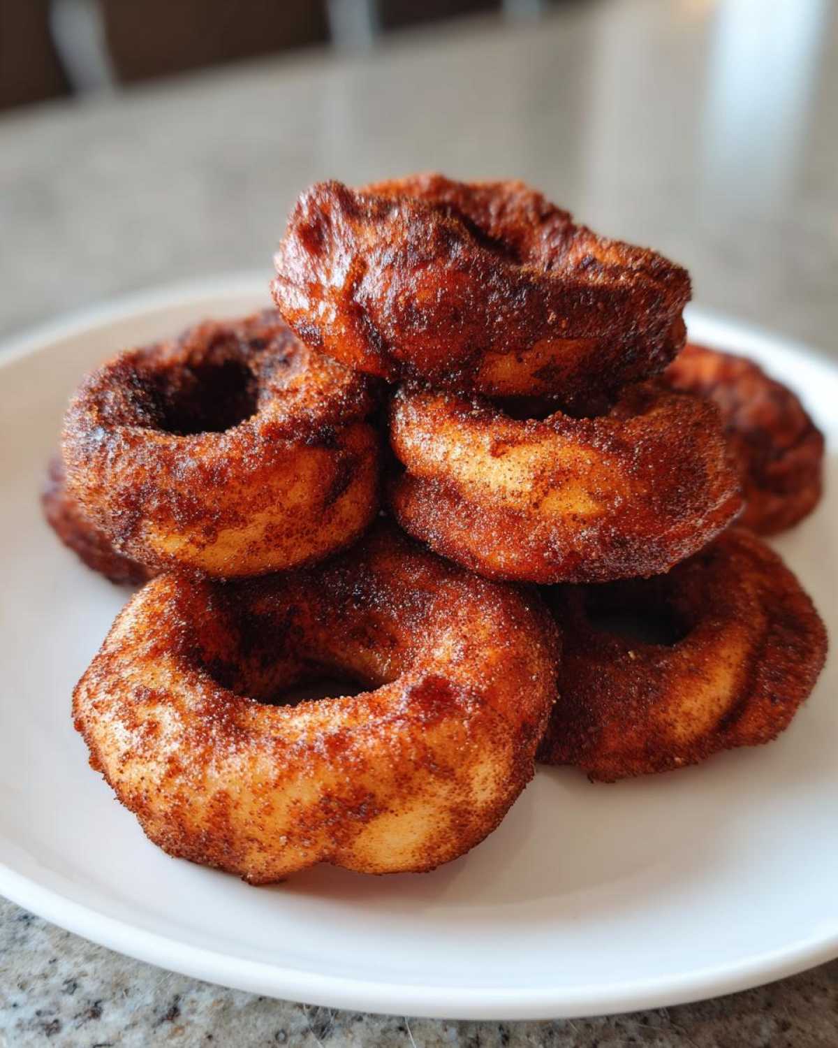 A stack of golden brown Air Fryer Cinnamon Apple Rings heavily coated in cinnamon sugar on a white plate.
