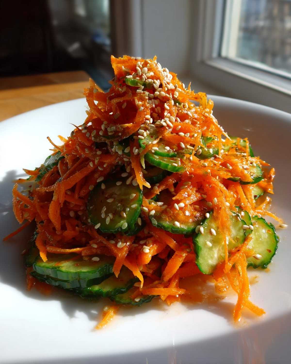 A mound of bright orange shredded carrots and sliced cucumbers tossed in a dressing and topped with sesame seeds, part of an Asian Carrot Cucumber Salad.