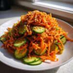 A mound of shredded carrots and sliced cucumbers tossed together, topped with sesame seeds, ready to serve as an Asian Carrot Cucumber Salad.