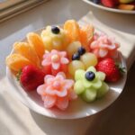 A white plate featuring a stunning Baby Bloom Fruit Platter with carved melon flowers, strawberries, orange segments, and blueberries.