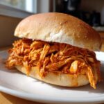 A close-up of a hearty Buffalo Chicken Crock Pot Sandwiches overflowing with saucy, shredded chicken on a soft bun.
