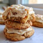 A stack of three Cozy Carrot Cake Cookies Cream, with the top cookie broken open showing the soft interior and thick cream cheese frosting.