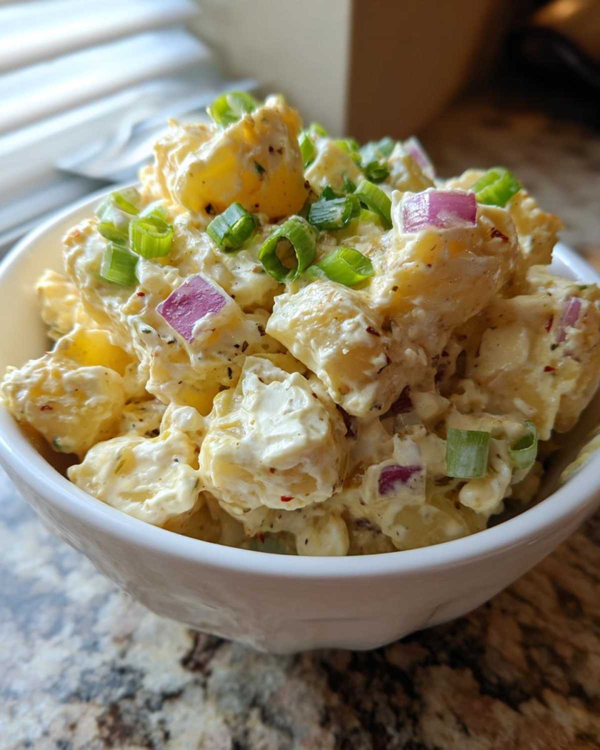 Close-up of creamy Jamaican Jerk Potato Salad in a white bowl, topped with chopped red onion and green onions.
