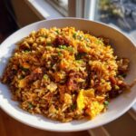A close-up of a white bowl filled with vibrant Creole Garlic Fried Rice mixed with chunks of sausage and scrambled egg.