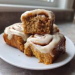 Close-up of three Easy Banana Bread Cinnamon Rolls stacked on a white plate, dripping with white cream cheese frosting.