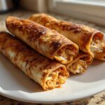 Four crispy, golden-brown Easy Homemade Chicken Taquitos piled on a white plate, showing shredded chicken filling.