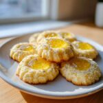 A stack of Easy Lemon Lavender Thumbprint Cookies with bright yellow centers and sprinkled lavender buds on a light gray plate.
