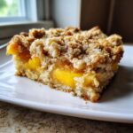 A close-up of a square slice of Easy Peach Cinnamon Oatmeal Crumb with visible layers of soft peaches and a thick, crumbly topping.