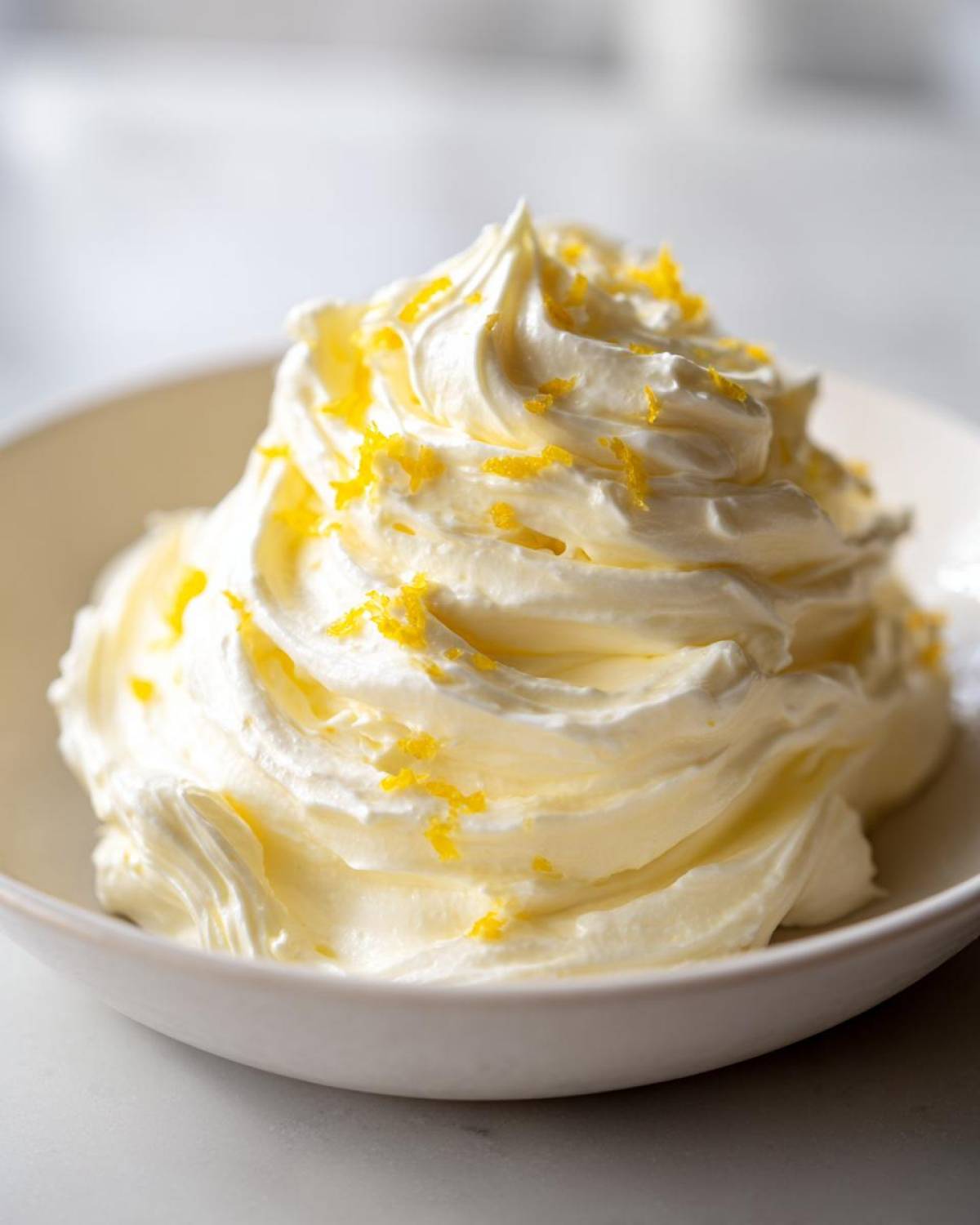 A beautiful swirl of Easy Soft Lemon Cream Cheese topped with bright yellow lemon zest in a white bowl.