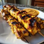 Close-up of golden yellow, charred Grilled Thai Chicken Skewers served on a white plate next to peanut sauce.