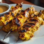 Four golden, grilled Thai chicken skewers with char marks, served next to a small bowl of peanut sauce.